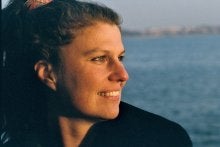 Photo of White woman looking towards right and smiling, sun on her face as she looks out at the water.
