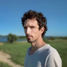 Man with brown curly hair and a slight mustache looks at the camera. He has pale skin and a white t-shirt, standing in a field with blue sky behind him