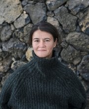 Photo of woman with brown hair and brown eyes and hair pulled back. Wearing black sweater in front of a stone wall 