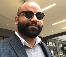 Photo of Ronald Baez, a Carribbean-American man with brown skin, a brown beard and wearing a gray suit with white button up shirt. He wears black sunglasses