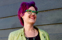 Producer Sara wears a green jacket with matching eyeglasses. She has pink hair and is smiling.