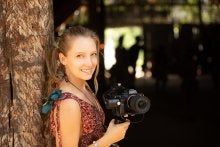 Photo of a blonde White woman leaning against a tree holding a camera